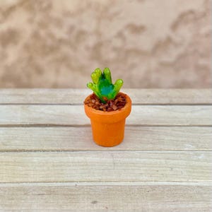 May include: A miniature orange terracotta pot containing a small, glossy green hand-shaped sculpture. The base of the sculpture is surrounded by dark brown, textured material that mimics soil, filling the top of the small, round container.