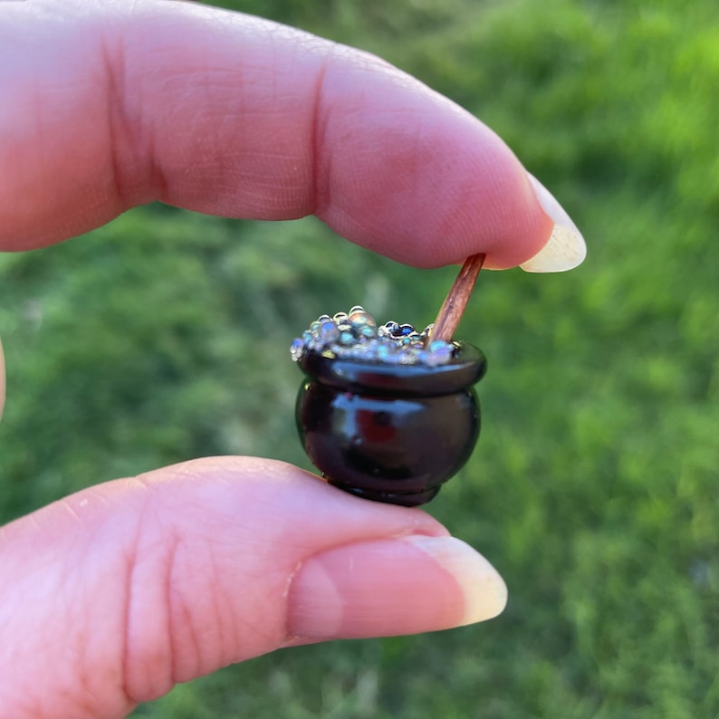 May include: A small black cauldron with a wooden stirring stick, filled with iridescent beads, resembling a bubbling potion.