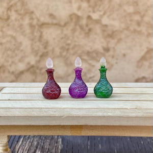 May include: Three miniature potion bottles in red, purple, and green, each featuring a textured surface with embossed eye motifs and a clear, pointed stopper. The bottles have a rounded base and a narrow neck, standing upright in a row.