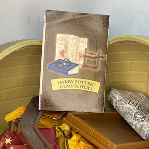 May include: A miniature Harry Potter Class Supplies book with illustrations of maps and glasses. The book is brown with a yellow banner. The book is in a wooden box with other miniature items.