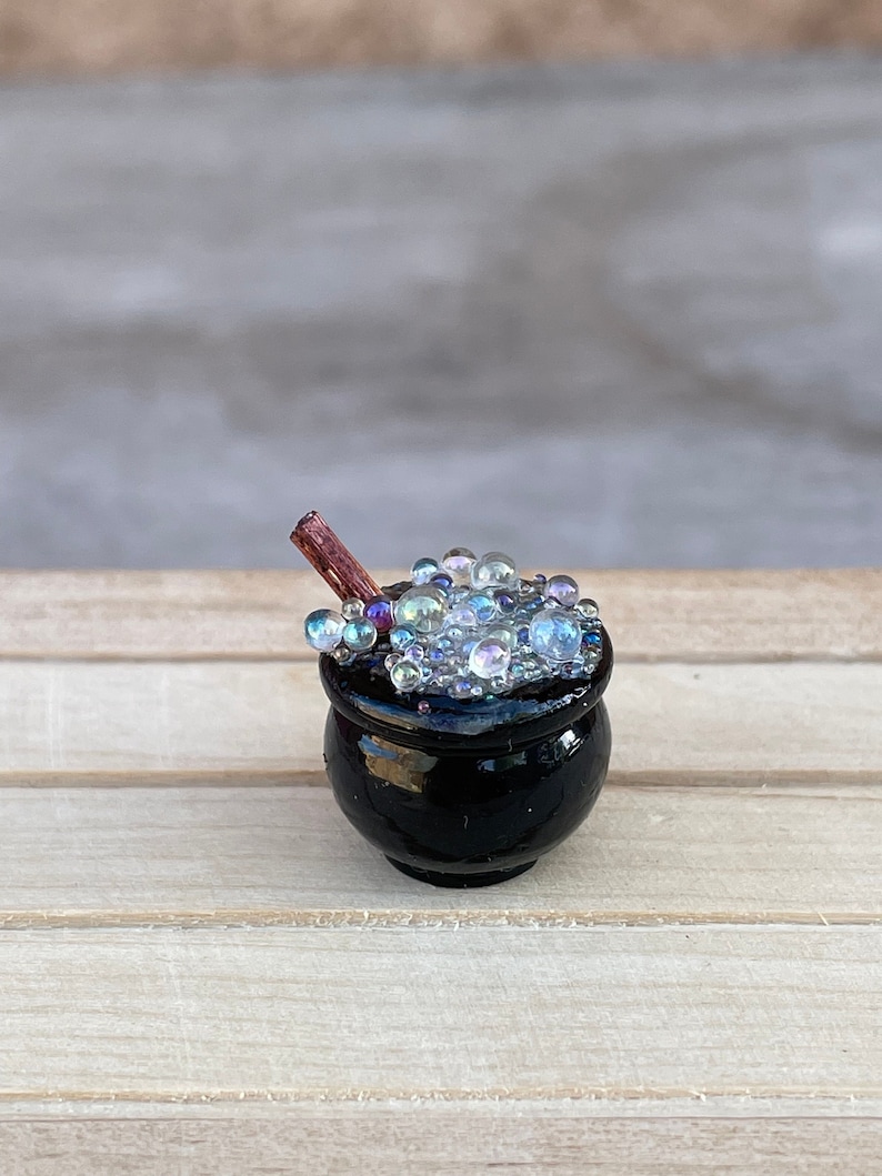 May include: A miniature black cauldron filled with iridescent bubbles and a brown wooden stirring stick.