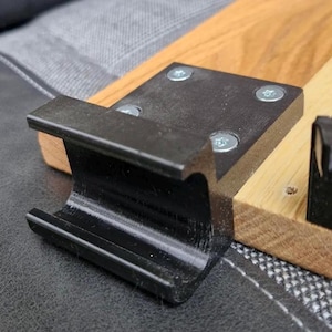 May include: A black metal bracket with a curved design, secured to a wooden base with four silver screws. The bracket has a smooth, glossy finish and is mounted on a light-coloured wood piece.