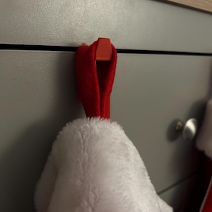 Könnte beinhalten: Ein roter Weihnachtsstrumpf hängt an einem roten Haken an einem grauen Schrank. Der Strumpf ist weiß und flauschig, mit einer roten Manschette. Der Haken ist an der Schublade des Schranks befestigt. Die Szene deutet auf ein festliches Urlaubsthema hin.