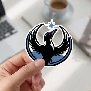 May include: A sticker featuring a stylized black bird with outstretched wings, a red eye, and a blue star above its head. The bird is set against a blue and white circular background. The sticker is held by a hand.