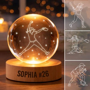 May include: A glowing softball-themed night light. The glass sphere features a white outline of a softball pitcher and star accents. The wooden base is engraved with "SOPHIA #26". Additional softball player illustrations are shown.