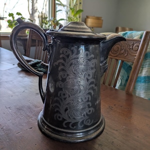 May include: Antique silver-plated coffee pot with a lid, handle, and spout. The pot features an ornate floral design etched into the surface. The pot is sitting on a wooden table.
