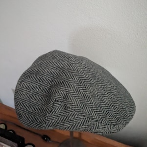 May include: A gray and black herringbone pattern newsboy cap. The cap is made of a woven fabric and has a rounded crown and a small, stiff brim. The cap is displayed on a metal stand.