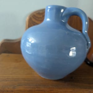 May include: A light blue ceramic pitcher with a rounded body and a curved handle. The pitcher is sitting on a wooden surface. The pitcher has a glossy finish and reflects light.