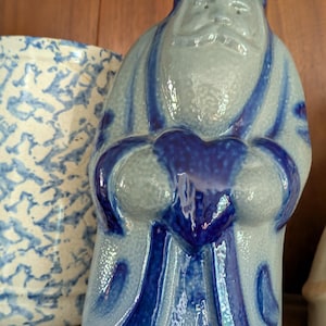 May include: A glazed ceramic figurine of a bearded figure wearing a hat and long robe, colored in light blue and dark blue. The figure holds a dark blue heart shape in front of the chest. The surface has a speckled texture and a glossy finish.
