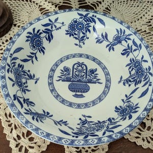 May include: A white ceramic serving bowl with a blue floral design. The bowl features a central oval with a basket of flowers and a detailed border. The rim is decorated with floral patterns. The bowl is placed on a cream-colored lace doily.