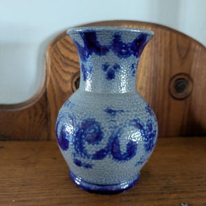 May include: A small, gray ceramic vase with a bulbous body and flared rim. The vase features a textured surface and is adorned with dark blue, swirling floral patterns. The vase is displayed on a wooden surface.