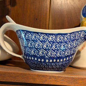 May include: A ceramic gravy boat featuring a white base with a deep blue hand-painted pattern. The design consists of repeating circular motifs and small dots arranged in horizontal bands around the exterior, complemented by a curved handle.