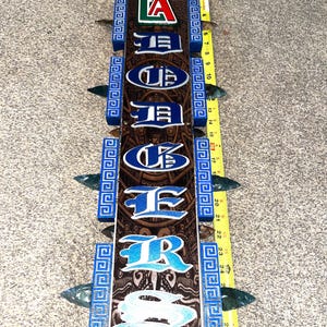May include: A wooden sign with the word "Lakers" in blue and white lettering. The sign has a brown background with Aztec-inspired designs and blue decorative borders. The sign is approximately 29 inches long.