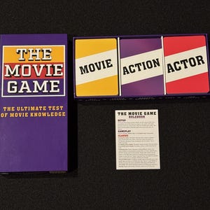 Może przedstawiać: Gra planszowa "The Movie Game" z fioletowym pudełkiem i kartami. Pudełko ma tytuł gry w białym i żółtym tekście. Karty są żółte, fioletowe i czerwone z białym tekstem. Dołączona jest również instrukcja.