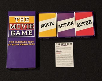 Puede incluir: Un juego de mesa llamado "The Movie Game" con una caja y cartas moradas. La caja tiene el título del juego en texto blanco y amarillo. Las cartas son amarillas, moradas y rojas con texto blanco. También se incluye un libro de reglas.