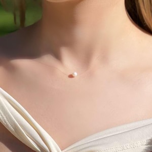 Könnte beinhalten: Eine zarte Halskette mit einer einzelnen Perle und einer fast unsichtbaren, transparenten Kette. Die Perle ist weiß und rund und hängt in der Mitte der Kette.