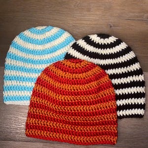 May include: Three crocheted beanies featuring horizontal stripes. One beanie has light blue and white stripes, another has black and white stripes, and the third beanie features alternating shades of vibrant orange and deep red yarn.