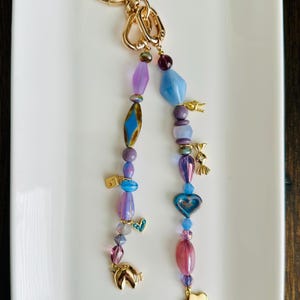 May include: A gold-coloured keyring with two beaded charms. The charms feature a variety of colourful beads in shades of purple, blue, and pink, along with gold-coloured charms shaped like hearts, clouds, and horseshoes. The keyring is on a white rectangular plate.