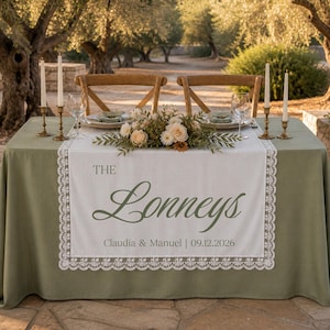 May include: A sage green tablecloth with a white lace-trimmed runner featuring the text THE Lonneys, Claudia & Manuel, and 09.12.2026. The table is set with two chairs, two wine glasses, two tall candles in gold holders, and a floral centerpiece.