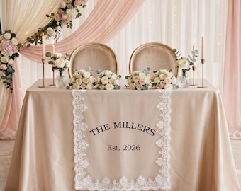 May include: A champagne-colored satin tablecloth featuring a white lace runner with the black text THE MILLERS Est. 2026. The table is set with two chairs, floral arrangements, and tall gold candlesticks against a backdrop of draped fabric.