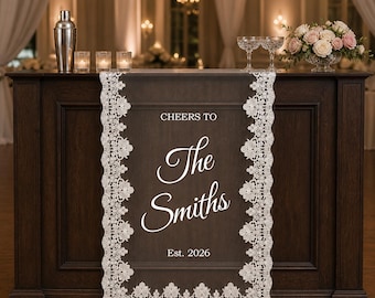 May include: A dark brown table runner featuring white lace trim along the edges. The center displays white text that reads CHEERS TO The Smiths Est. 2026. The runner is draped over a dark wooden bar counter, creating a decorative accent for events.