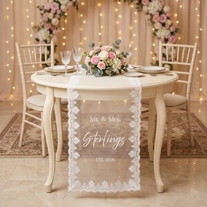 Puede incluir: Una mesa redonda color crema preparada para una recepci&oacute;n de boda. Un camino de mesa de encaje blanco transparente con el texto "Mr. & Mrs. Sterlings EST. 2026" est&aacute; centrado en la mesa. La mesa est&aacute; puesta con platos, cubiertos y copas de vino, y un arreglo floral.