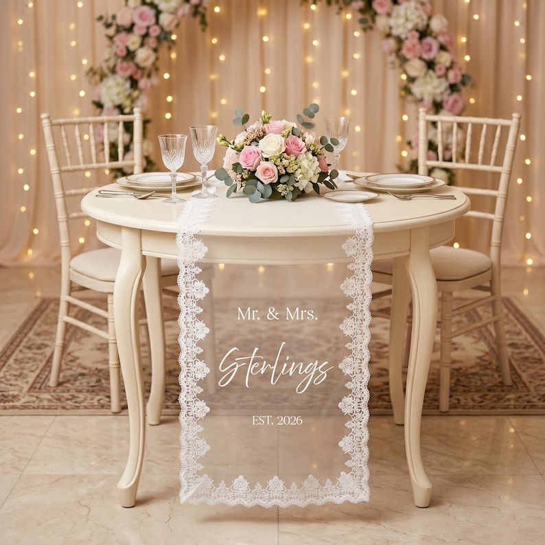 Puede incluir: Una mesa redonda color crema preparada para una recepci&oacute;n de boda. Un camino de mesa de encaje blanco transparente con el texto "Mr. & Mrs. Sterlings EST. 2026" est&aacute; centrado en la mesa. La mesa est&aacute; puesta con platos, cubiertos y copas de vino, y un arreglo floral.