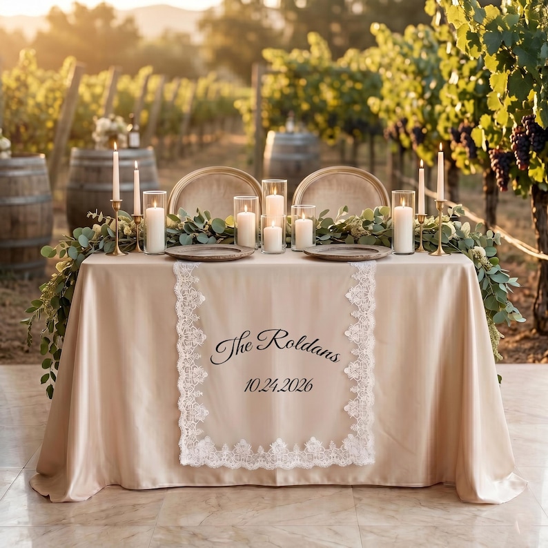 Puede incluir: Una mesa de recepci&oacute;n de bodas al aire libre con un mantel color crema y ribete de encaje. La mesa est&aacute; decorada con velas, vegetaci&oacute;n y cubiertos. El texto "The Roldans 10.24.2026" est&aacute; impreso en el mantel.