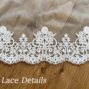 Puede incluir: Primer plano de un ribete de encaje blanco con intrincados dise&ntilde;os florales y de volutas. El encaje se coloca sobre una tela transparente y un fondo de madera. El texto "Lace Details" est&aacute; en la parte inferior.