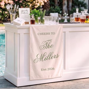 Könnte beinhalten: Ein cremefarbenes Stoffbanner, das über einer weißen Bar hängt und den grünen Text "CHEERS TO The Millers Est.2026" in einer Mischung aus Serif- und Schreibschriften zeigt. Das Banner hängt vertikal und zeigt den Familiennamen und das Datum.
