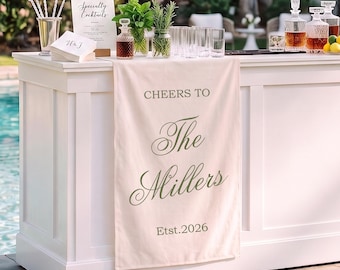May include: A cream-colored fabric table runner draped over a white bar, featuring the dark green printed text "CHEERS TO The Millers Est.2026". The runner hangs vertically, displaying elegant script and serif typography on a smooth, rectangular textile surface.