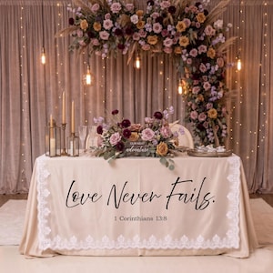 Puede incluir: Una mesa decorada con un mantel crema con encaje y la frase "Love Never Fails". Arreglos florales, velas y un arco floral adornan la mesa, creando un ambiente rom&aacute;ntico.