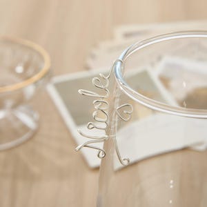May include: Silver wire name charm with the name "Nicole" and a small heart charm. The charm is attached to a clear glass cup.