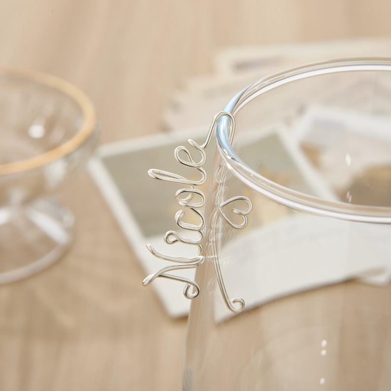 May include: Silver wire name charm with the name "Nicole" and a small heart charm. The charm is attached to a clear glass cup.