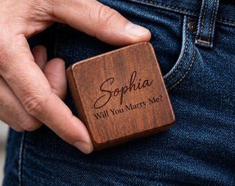 May include: A small, square, dark brown wooden box with a smooth finish. The lid is engraved with the name Sophia in a cursive script, followed by the text Will You Marry Me? in a clean, sans-serif font, centered on the wood surface.