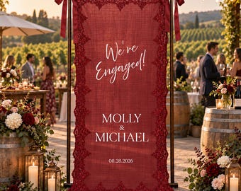 May include: A vertical deep red fabric banner suspended from a dark metal frame, featuring ornate lace trim along the edges. The text reads We're Engaged! MOLLY & MICHAEL 08.28.2026 in white lettering. The banner is secured at the top with red ribbons.