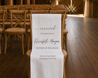 May include: A white fabric memorial sign draped over a wooden chair. The text reads: reserved, In loving memory of, Elizabeth Morgan, MOTHER OF THE GROOM, Forever in our hearts, always part of our day. The sign features black lettering.