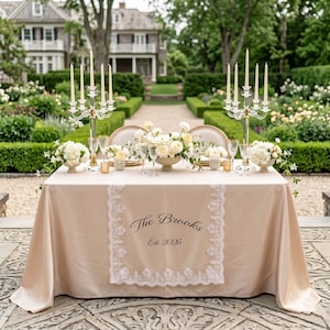 Puede incluir: Una mesa bellamente decorada para una recepci&oacute;n de boda, con un mantel color champ&aacute;n y un camino de mesa de encaje. La mesa est&aacute; adornada con arreglos florales, velas y cristaler&iacute;a. El texto "The Brooks Est. 2026" est&aacute; impreso en el camino de mesa.
