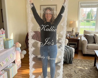 May include: A white lace baby shower decoration with the text "WELCOME TO Kallie Jo's Baby Shower". The lace has a delicate floral design around the edges. The decoration is held up by a person.