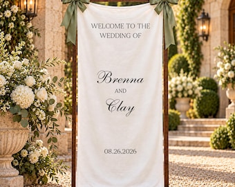 May include: A vertical white fabric wedding welcome sign suspended from a dark wooden frame. The sign features black text reading Welcome to the wedding of Brenna and Clay, dated 08.26.2026. Two sage green satin bows are tied to the top corners.