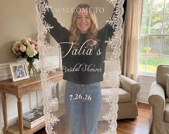 May include: A rectangular white lace veil featuring intricate floral embroidery along the edges. The center of the sheer fabric displays the white printed text: WELCOME TO Talia's Bridal Shower 7.26.26. The veil is held vertically by its top corners.