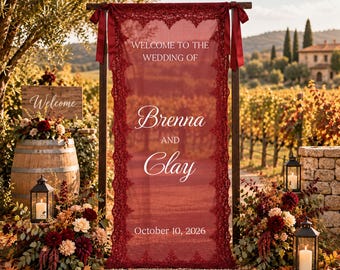 May include: A vertical deep red fabric wedding sign featuring white text that reads Welcome to the wedding of Brenna and Clay October 10, 2026. The banner is framed by intricate dark red lace borders and is suspended from a wooden frame with ribbons.