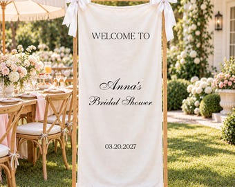 May include: A vertical cream-colored fabric banner suspended on a wooden frame with white ribbon bows at the top corners. The text reads: WELCOME TO, Anna's Bridal Shower, and 03.20.2027 in black lettering. The banner is displayed outdoors.
