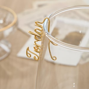 May include: Gold wire name card holder with the name "Jordan" written on it. The holder is attached to a clear glass cup.