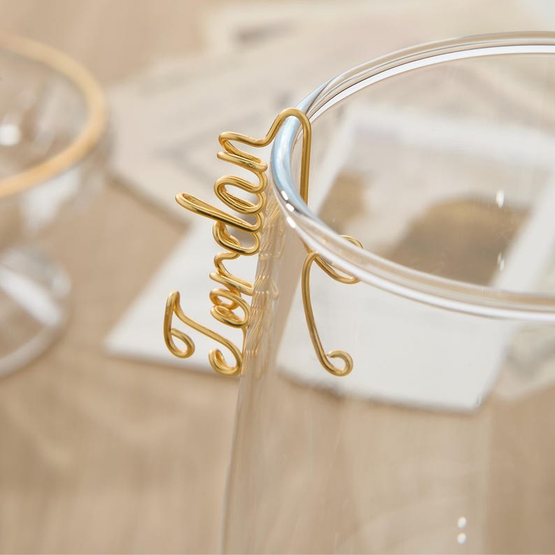 May include: Gold wire name card holder with the name "Jordan" written on it. The holder is attached to a clear glass cup.