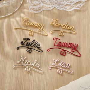 May include: Six personalized wire name hangers with heart accents. The names are Tammy, Jordan, Julie, Tammy, Nicole, and Lisa. The hangers are in gold, red, black, silver, and pink.