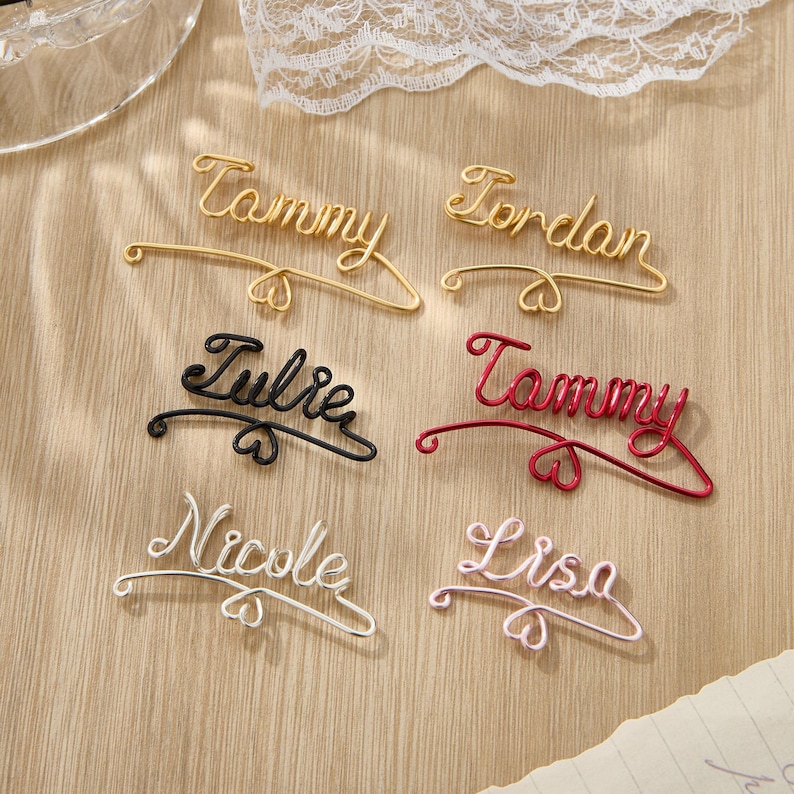 May include: Six personalized wire name hangers with heart accents. The names are Tammy, Jordan, Julie, Tammy, Nicole, and Lisa. The hangers are in gold, red, black, silver, and pink.