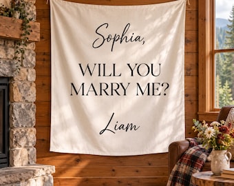 May include: A rectangular off-white fabric wall hanging featuring black printed text that reads: Sophia, WILL YOU MARRY ME? Liam. The banner is suspended from a horizontal rod against a wooden wall, displaying a proposal message in script and serif fonts.