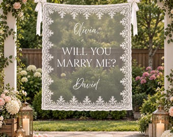 May include: A rectangular white lace banner featuring the text Olivia, WILL YOU MARRY ME? David. The banner is suspended by white ribbons from a wooden structure, showcasing a detailed floral lace border pattern on a sheer fabric background.