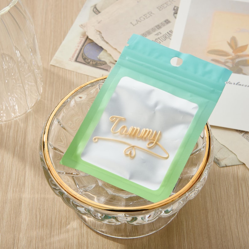 May include: A clear plastic bag with a green and blue gradient design. The bag contains a gold-colored metal nameplate with the word "Tammy" written on it.
